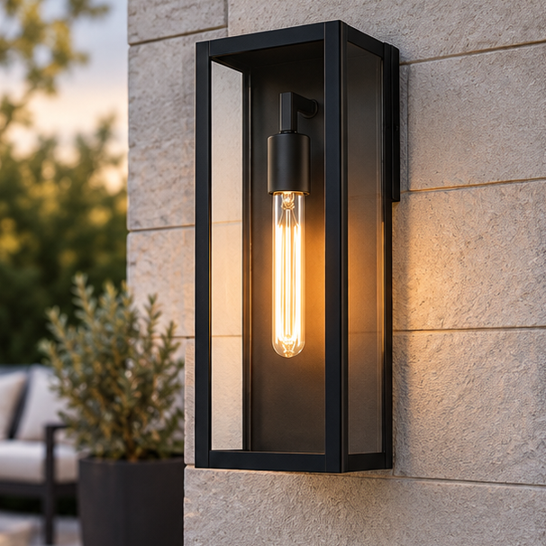 Black outdoor wall lantern with clear glass on a light stone block wall with a potted plant and patio seating behind.