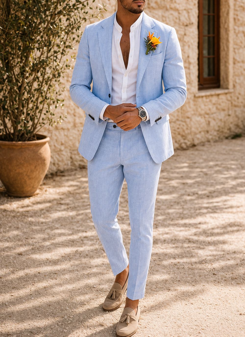 Model in the light blue suit with white shirt and wristwatch, hands clasped in front, standing on a gravel path.