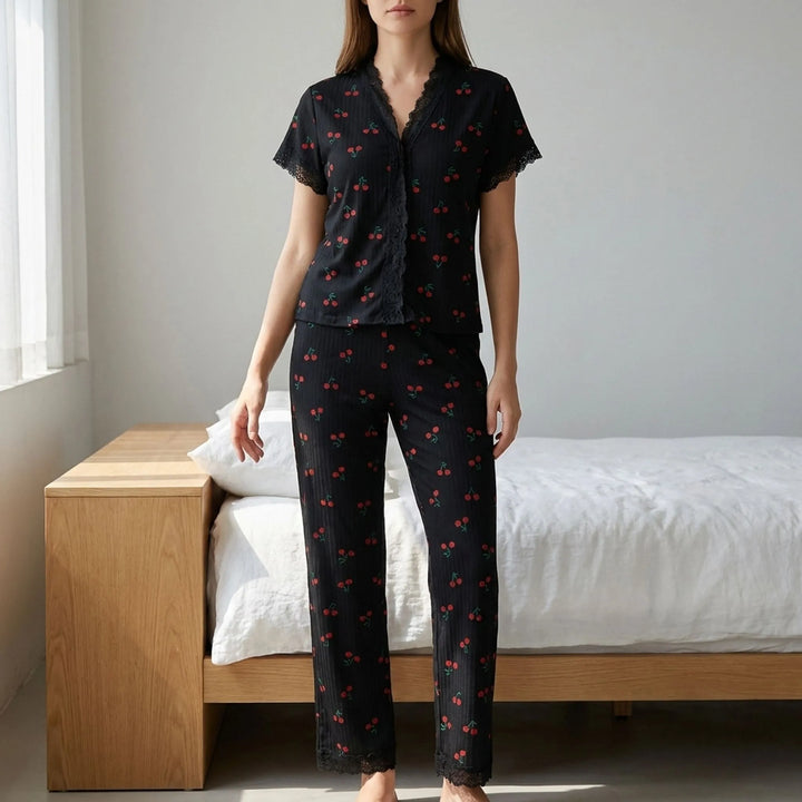 Model standing in a bedroom wearing the black ribbed short-sleeve pajama top with lace trim and matching cherry-print pants. 1