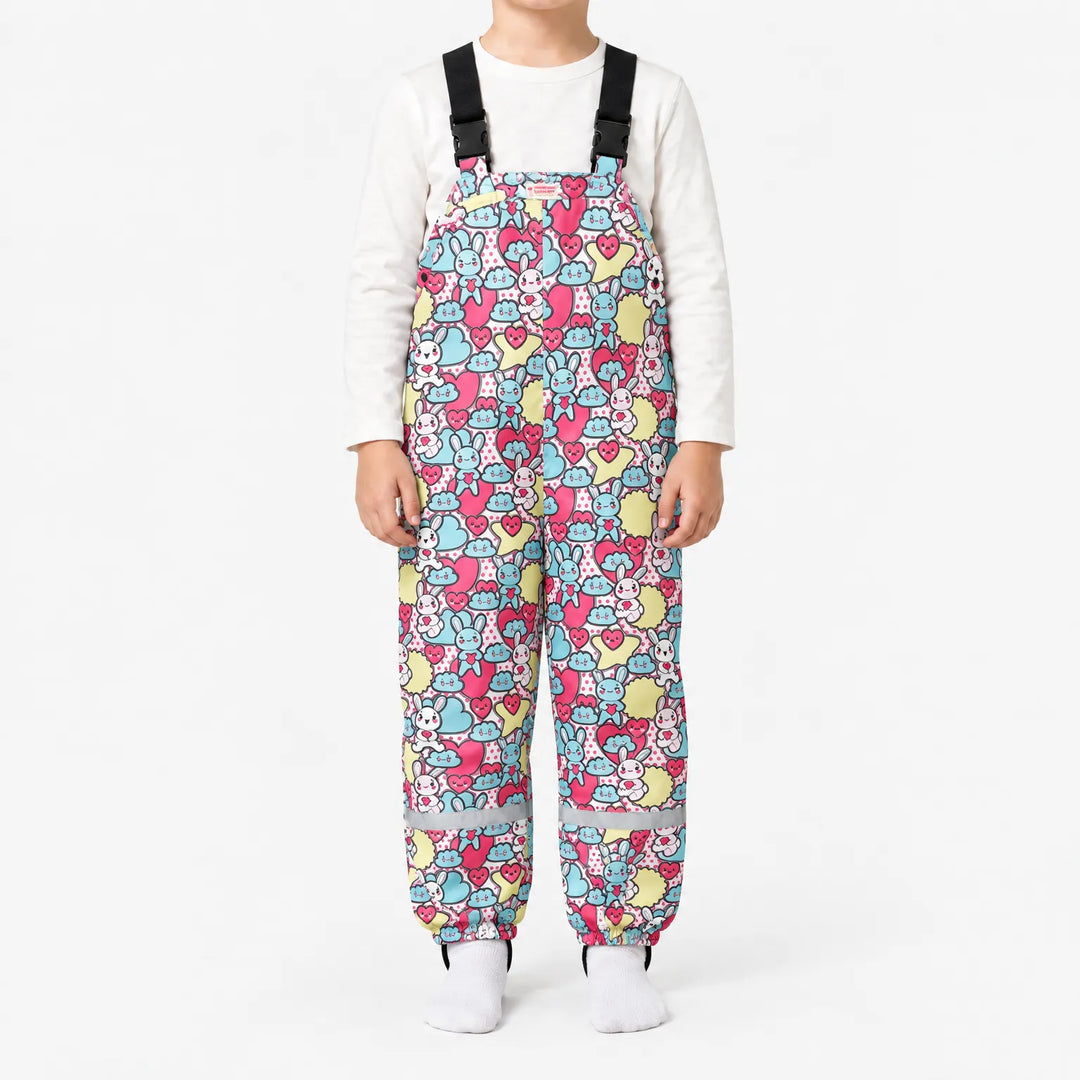 Child wearing dungarees with a busy pattern of cartoon bunnies, hearts and clouds in pink, blue and yellow, with reflective ankle bands. 15