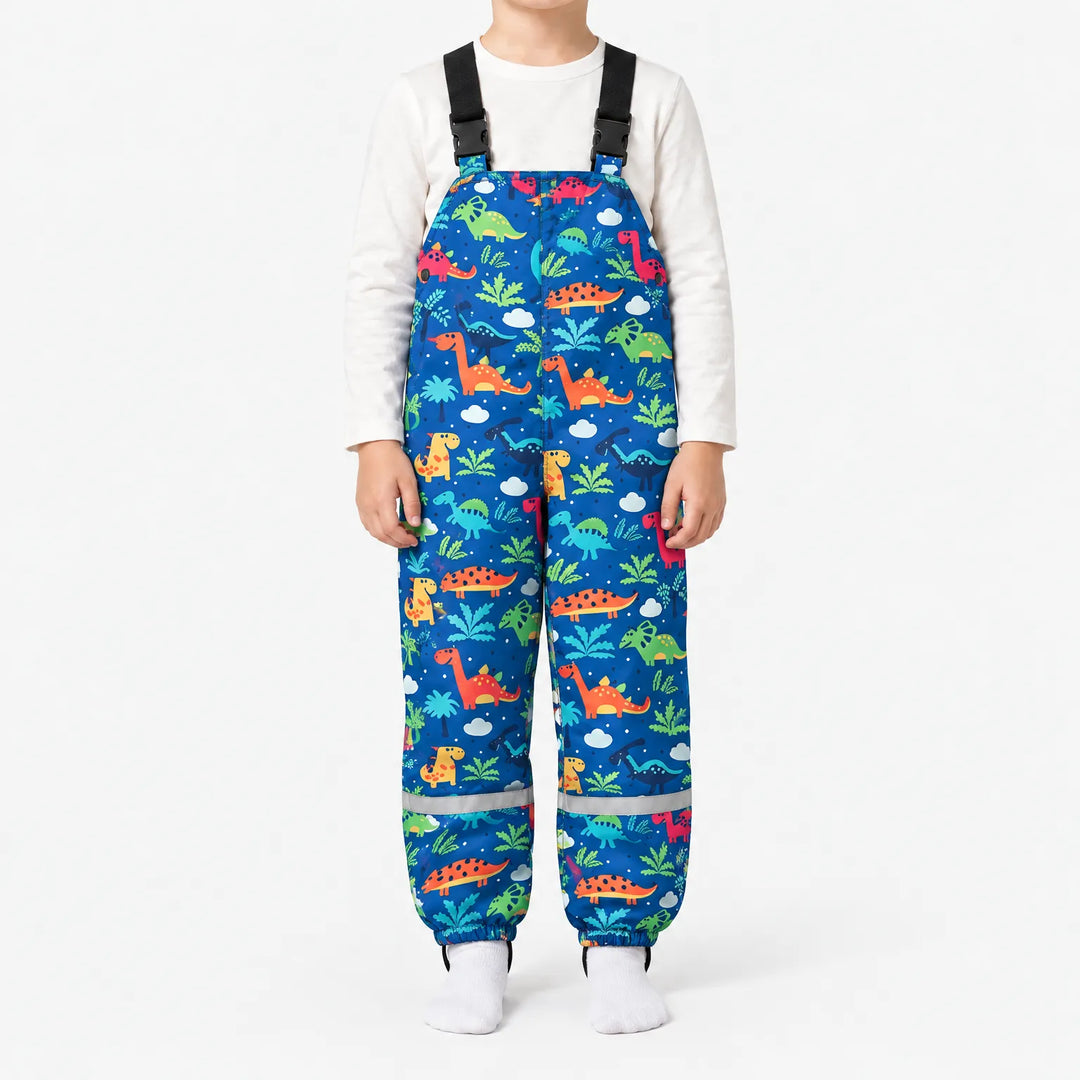 Child wearing royal blue dungarees printed with colorful dinosaurs, palm trees and clouds, with black buckled straps and reflective ankle bands. 16