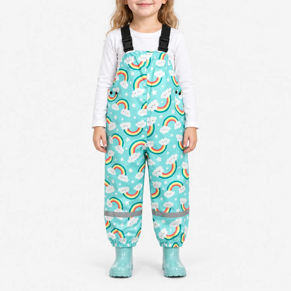 Smiling girl wearing turquoise dungarees printed with smiling cloud rainbows and stars, with matching boots and a reflective band at each ankle. 2