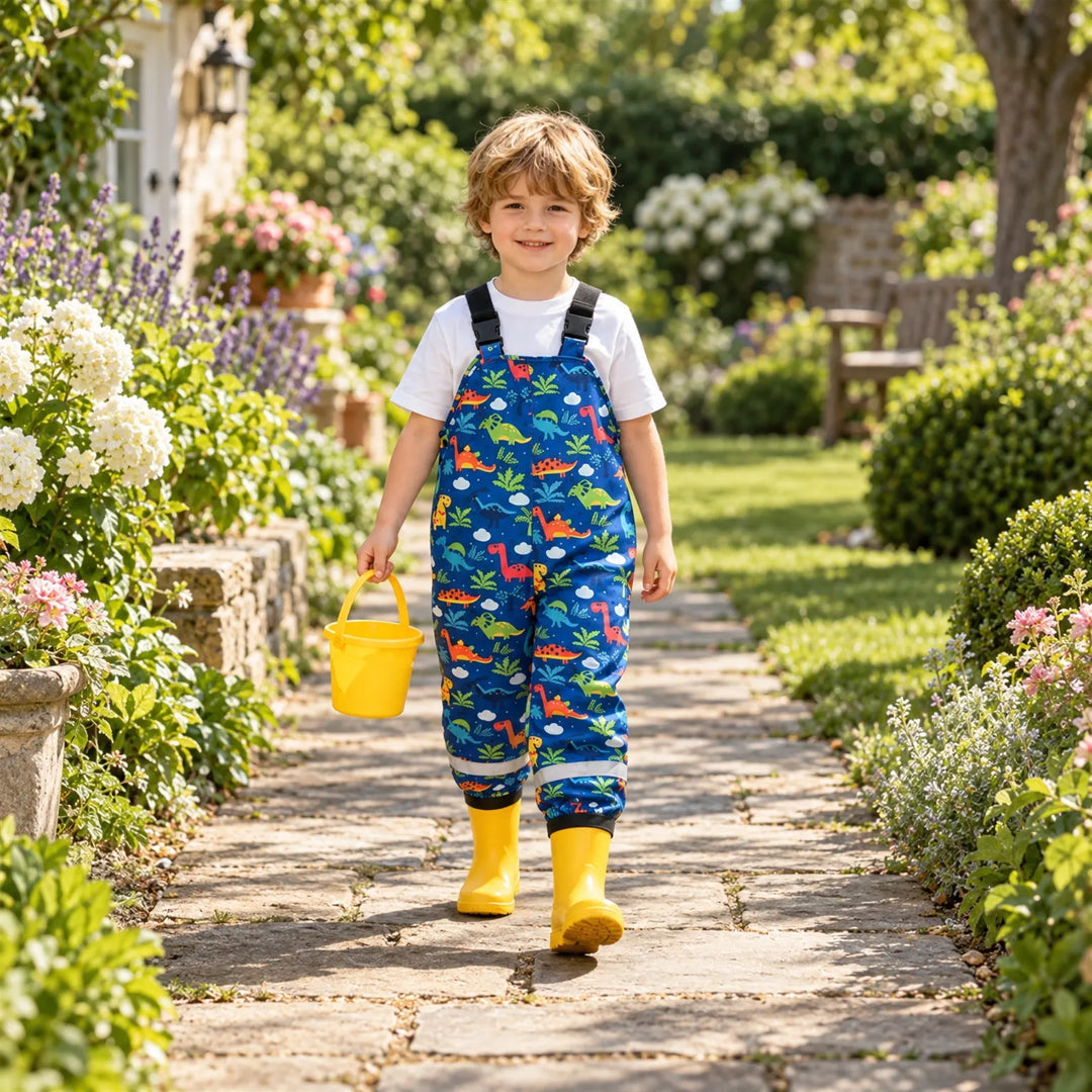 Smiling boy in blue dinosaur-print dungarees and yellow rain boots walking along a sunny garden path while carrying a yellow bucket. 3