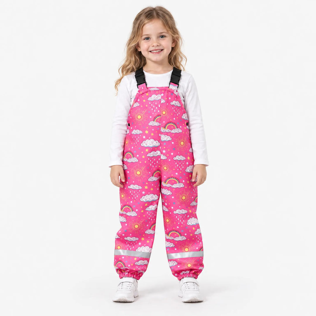 Smiling girl in pink dungarees printed with clouds, rainbows, suns and stars, worn over a white top with white sneakers, front view. 4