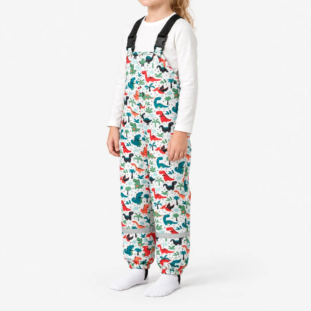 Side view of a child wearing white dungarees with colorful dinosaurs and palm trees, showing the black buckled straps and reflective ankle band. 5