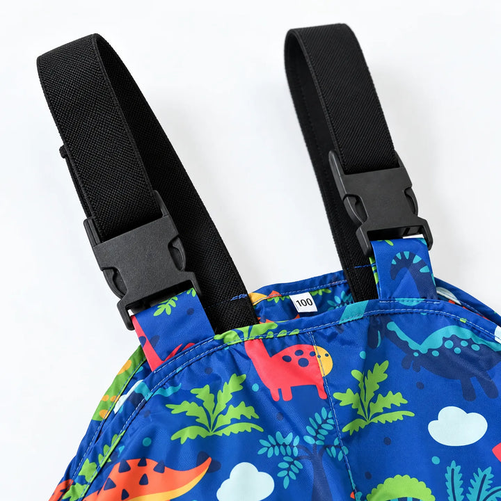 Close-up of the black adjustable shoulder straps with plastic buckles on blue dinosaur-print dungarees, with a small size label inside the bib. 6