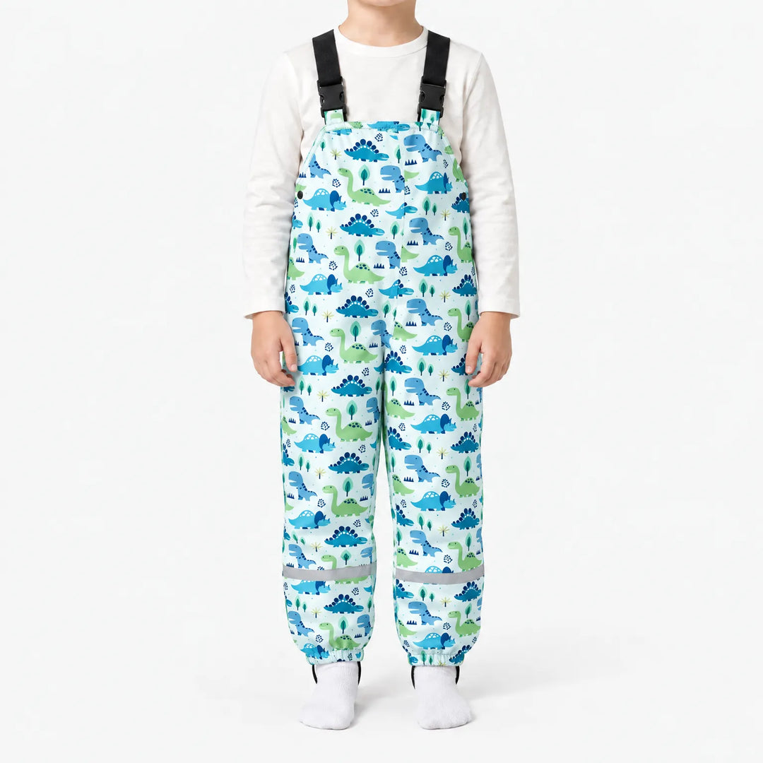 Child wearing light dungarees printed with blue and green cartoon dinosaurs and little trees, with black buckled straps and reflective leg bands. 9