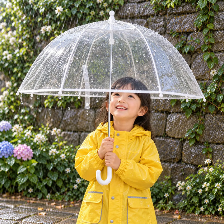 Smiling girl in a yellow raincoat holds the clear dome umbrella with raindrops on it, standing by a flowering garden wall. 1