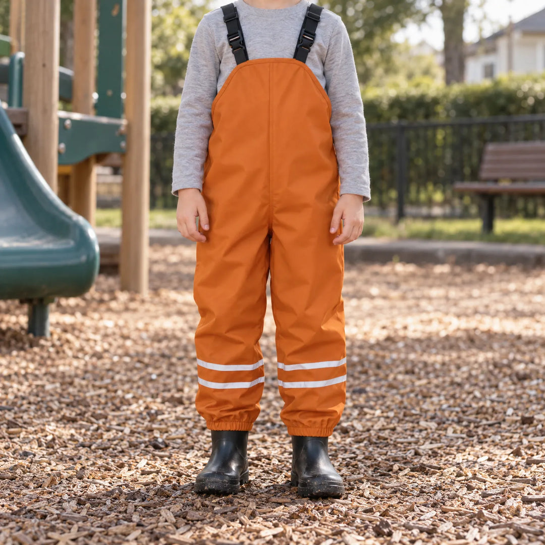 Child wearing orange waterproof overalls over a gray long-sleeve shirt with black rain boots, standing on playground wood chips. 3