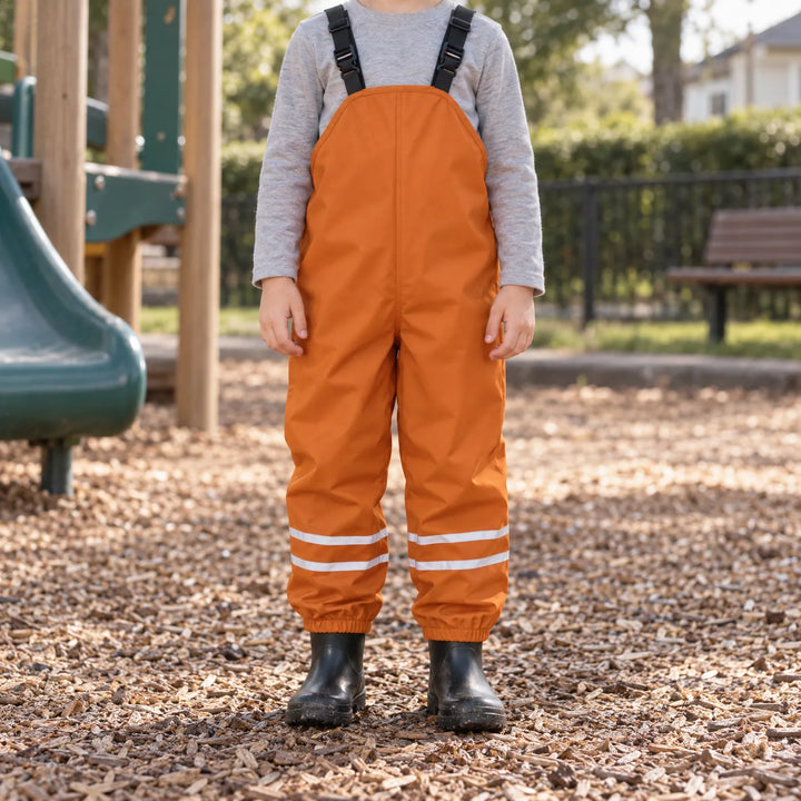 Child wearing orange waterproof overalls over a gray long-sleeve shirt with black rain boots, standing on playground wood chips. 3