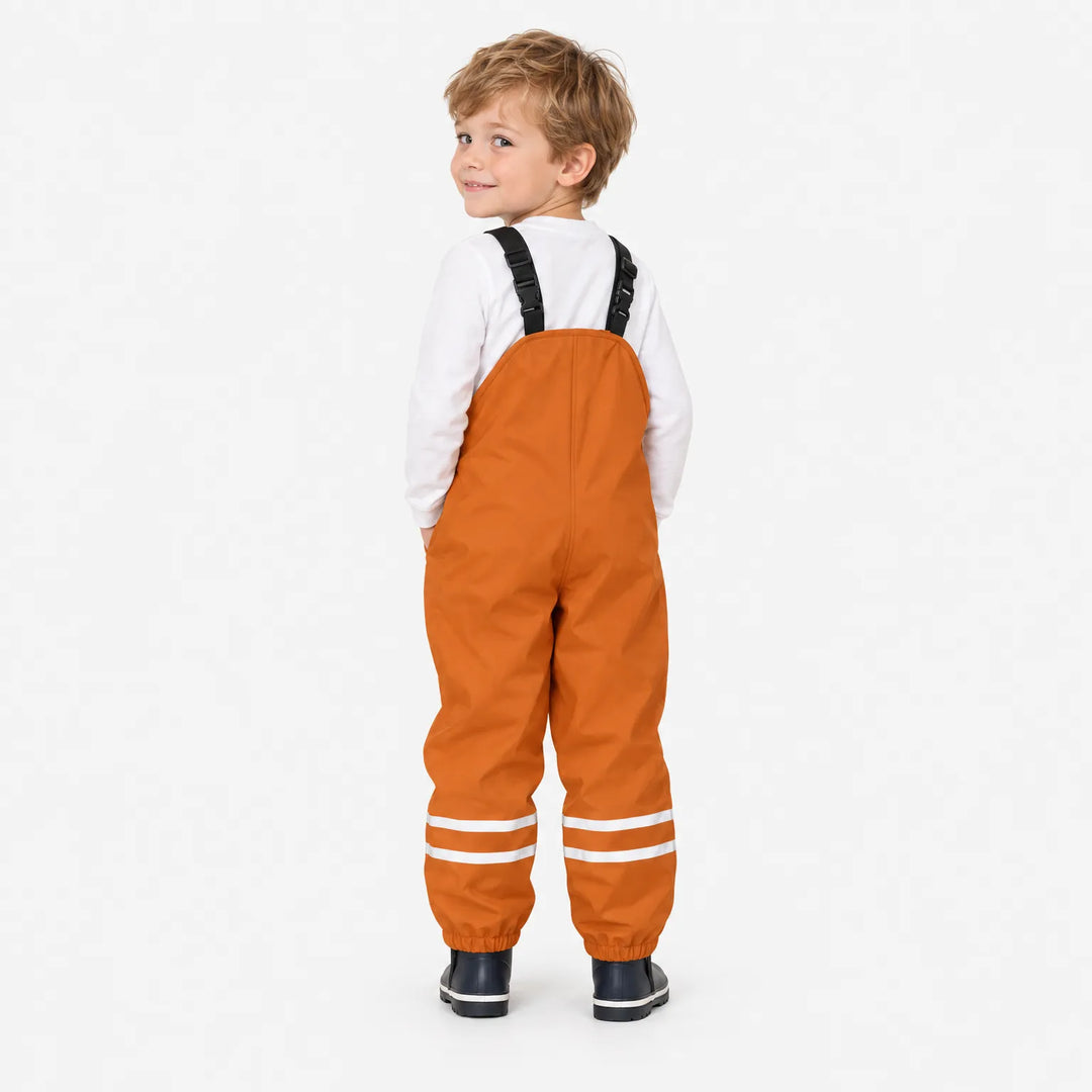 Back view of a smiling boy looking over his shoulder in orange waterproof overalls with reflective ankle stripes and navy boots. 4