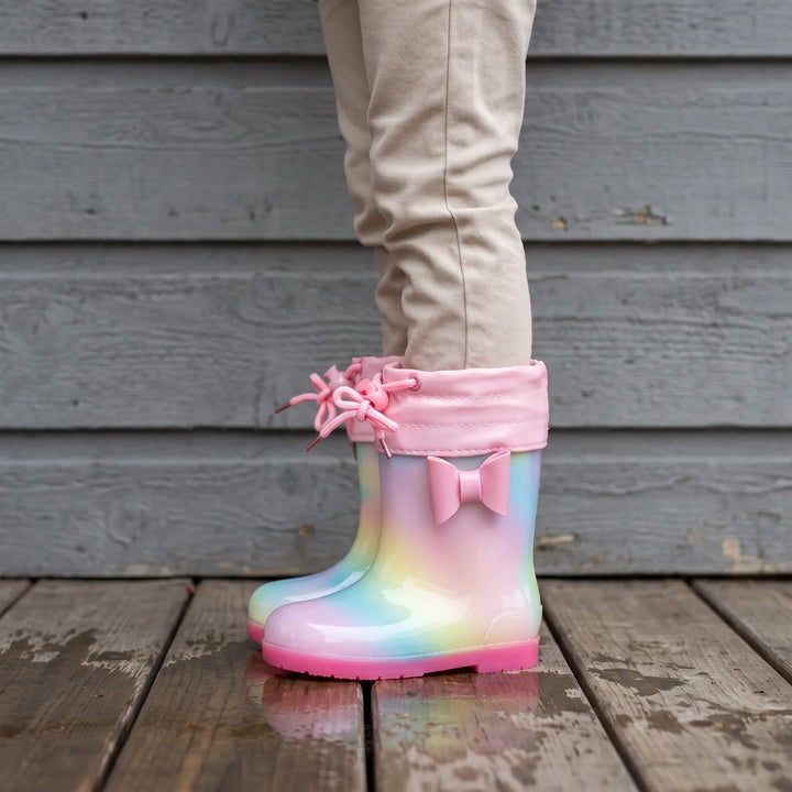 Child in beige pants wearing rainbow rain boots with a pink drawstring cuff and bow, standing on a wooden deck. 3