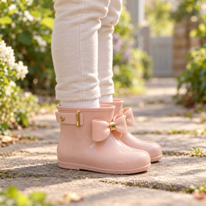 Child in cream leggings and white socks wears glossy blush pink rain boots with bows, standing on a sunny garden path. 1