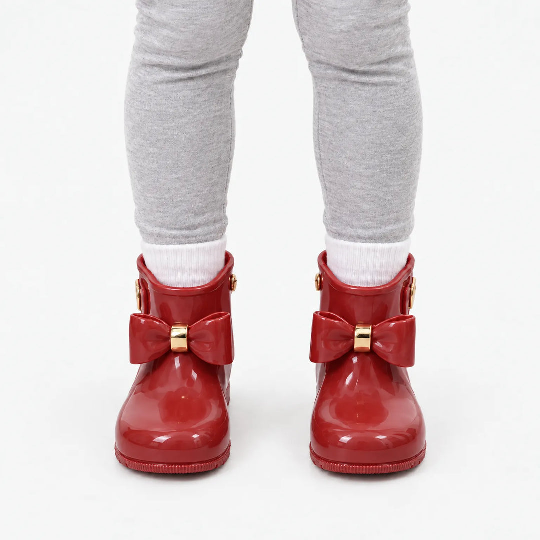 Front view of a child in gray leggings and white socks wearing glossy red rain boots with bows and gold accents. 10