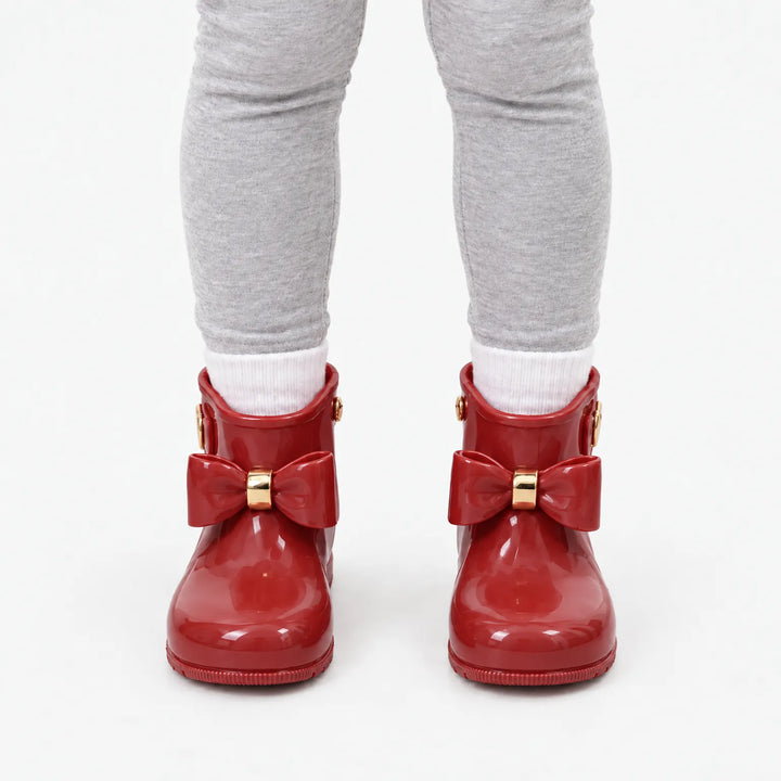 Front view of a child in gray leggings and white socks wearing glossy red rain boots with bows and gold accents. 10