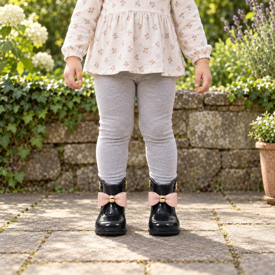 Toddler in a floral top and gray leggings stands in a garden wearing glossy black boots with blush pink bows. 3