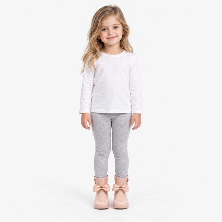 Smiling girl in a white long-sleeve top and gray leggings wears blush pink rain boots with bows, full-length studio shot. 4