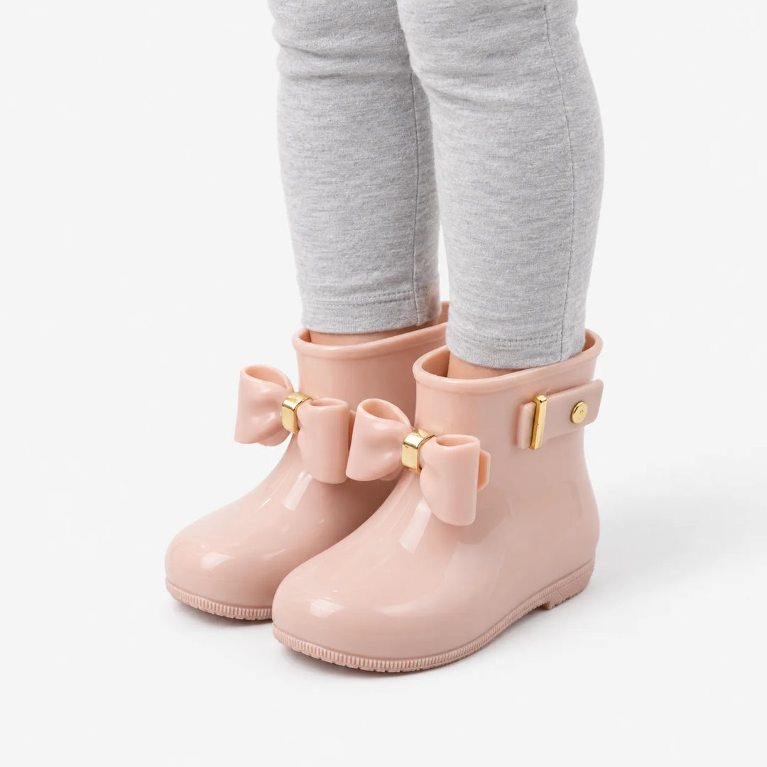 Angled side view of a child's legs in gray leggings wearing blush pink rain boots with bows and a buckle strap. 5