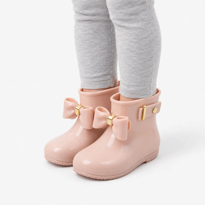 Angled side view of a child's legs in gray leggings wearing blush pink rain boots with bows and a buckle strap. 5