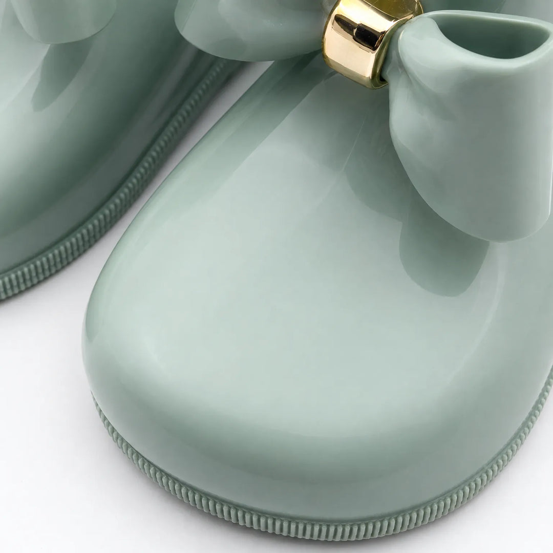 Close-up of a glossy sage green boot toe, showing the smooth finish, ridged sole edge and bow with gold ring. 6