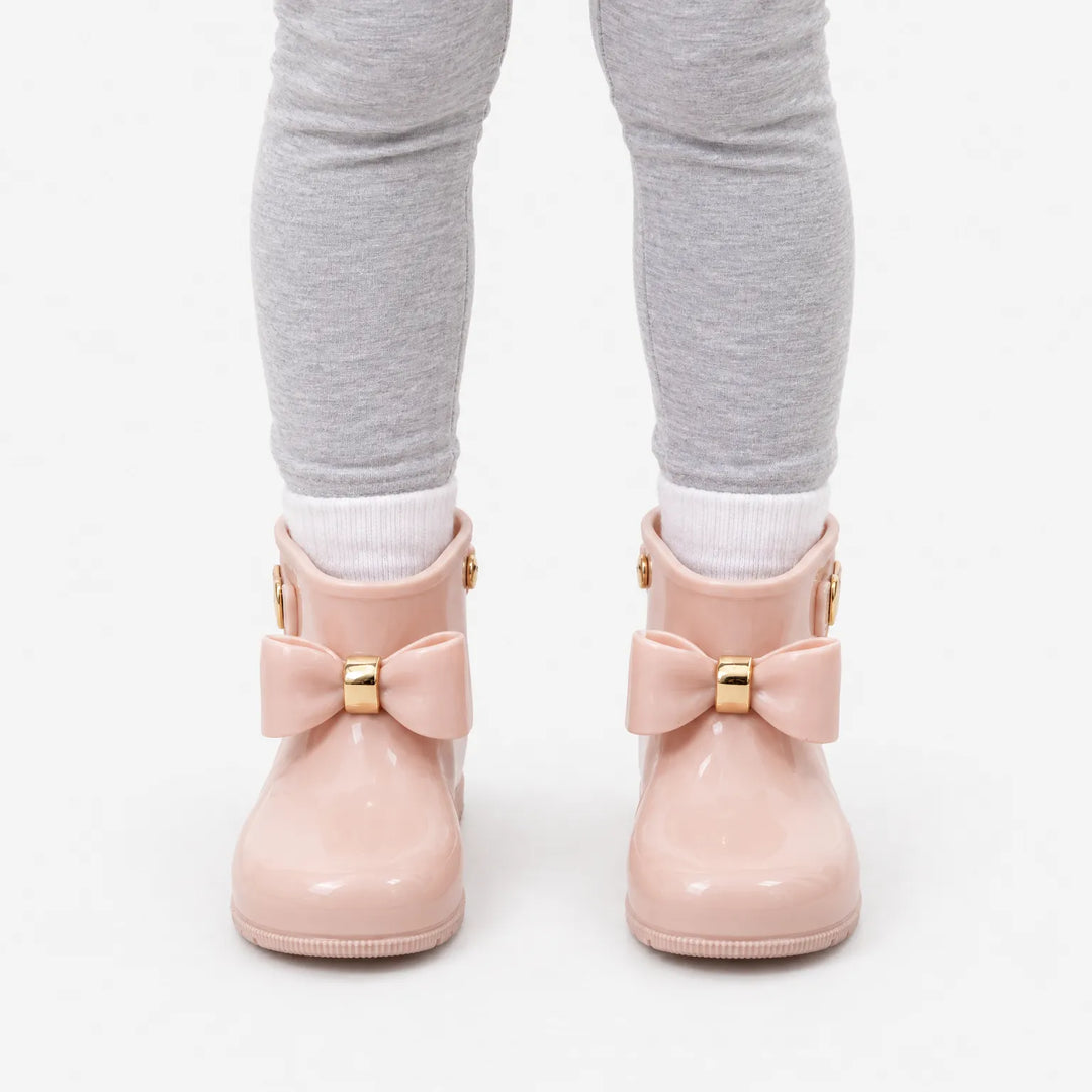 Front view of a child in gray leggings and white socks wearing blush pink rain boots with bows on a white background. 7