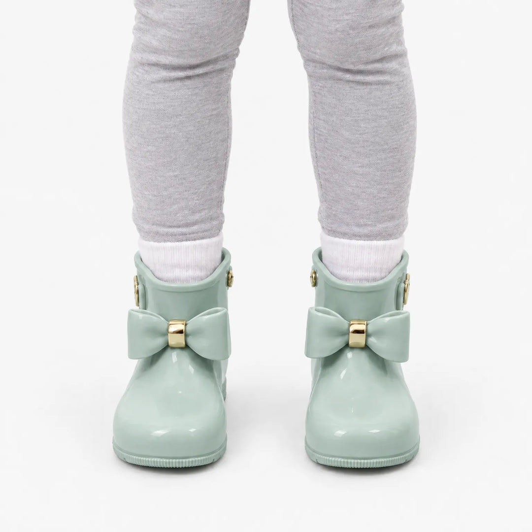 Front view of a child in gray leggings and white socks wearing sage green rain boots with bows and gold rings. 8