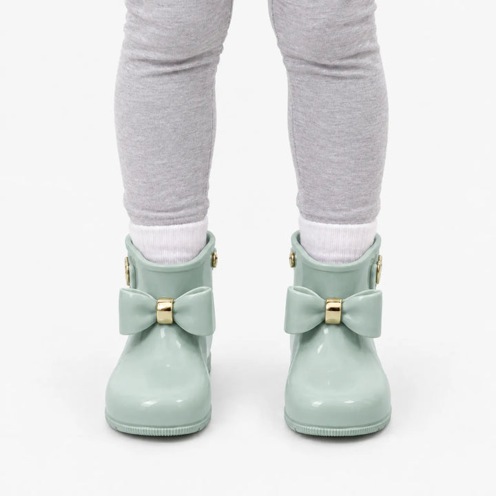 Front view of a child in gray leggings and white socks wearing sage green rain boots with bows and gold rings. 8