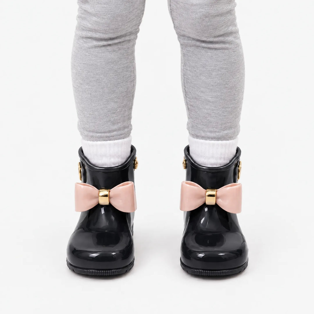 Front view of a child in gray leggings and white socks wearing glossy black rain boots with blush pink bows. 9