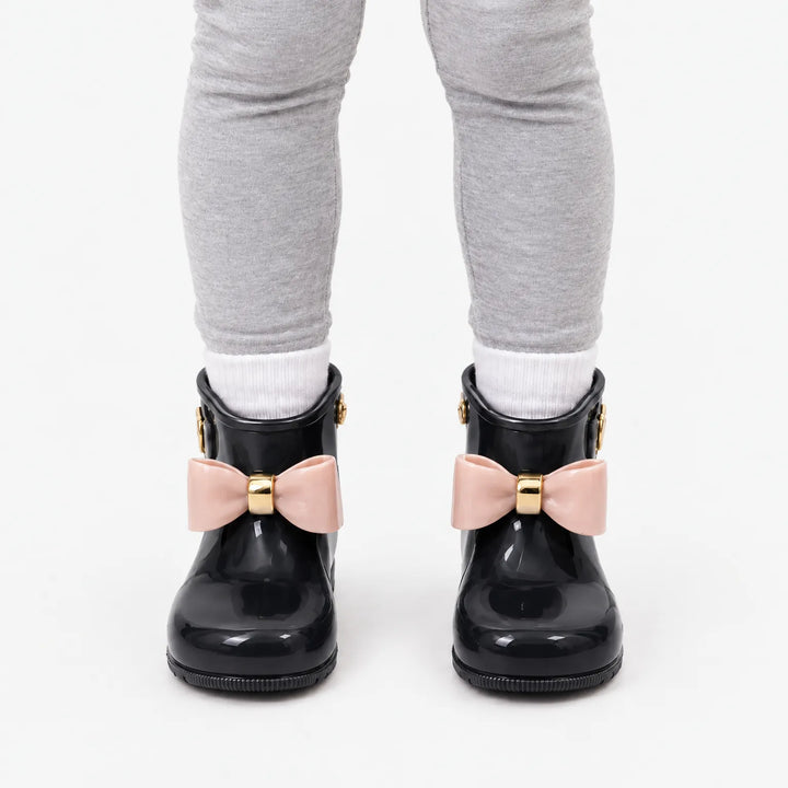 Front view of a child in gray leggings and white socks wearing glossy black rain boots with blush pink bows. 9