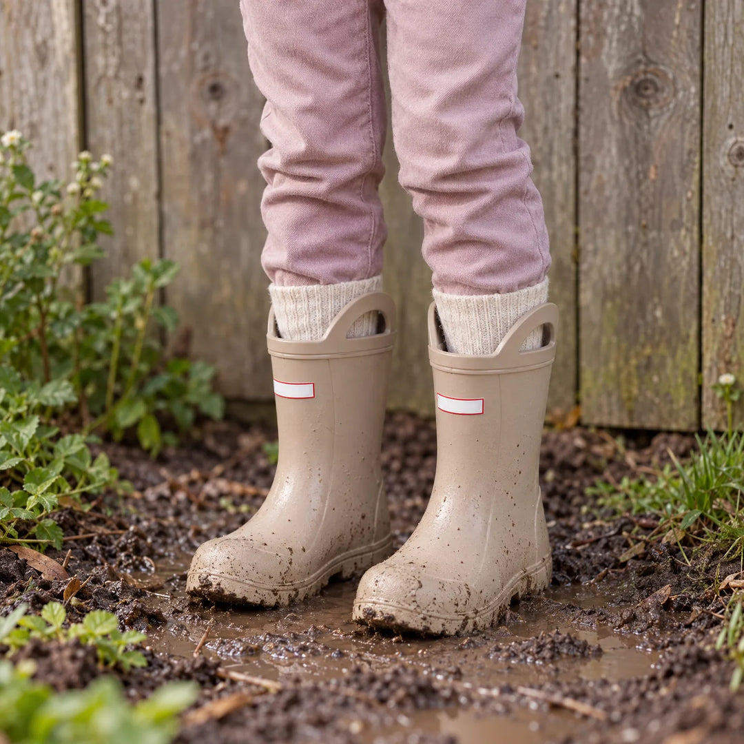 Child in beige rain boots with cream wool socks and pink pants standing in mud beside a wooden fence. 3
