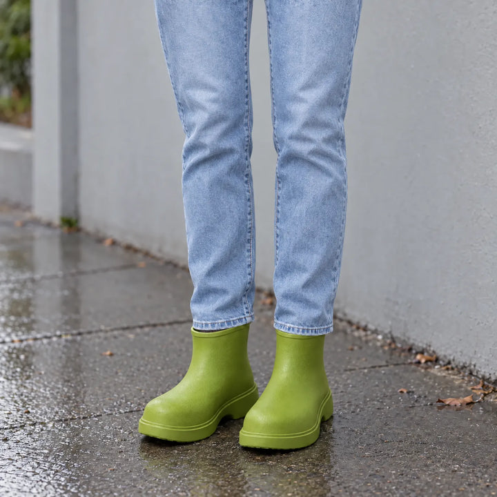 Green ankle boots worn with light-wash jeans on a wet city sidewalk, shown from the knees down. 1