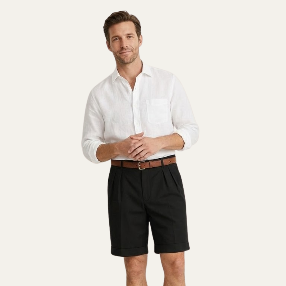Man in black pleated cuffed shorts, white linen shirt with chest pocket and brown belt, hands clasped in front.