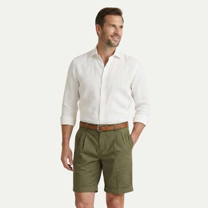 Man in olive green pleated cuffed shorts, white linen shirt and brown belt, looking aside with one hand in pocket.