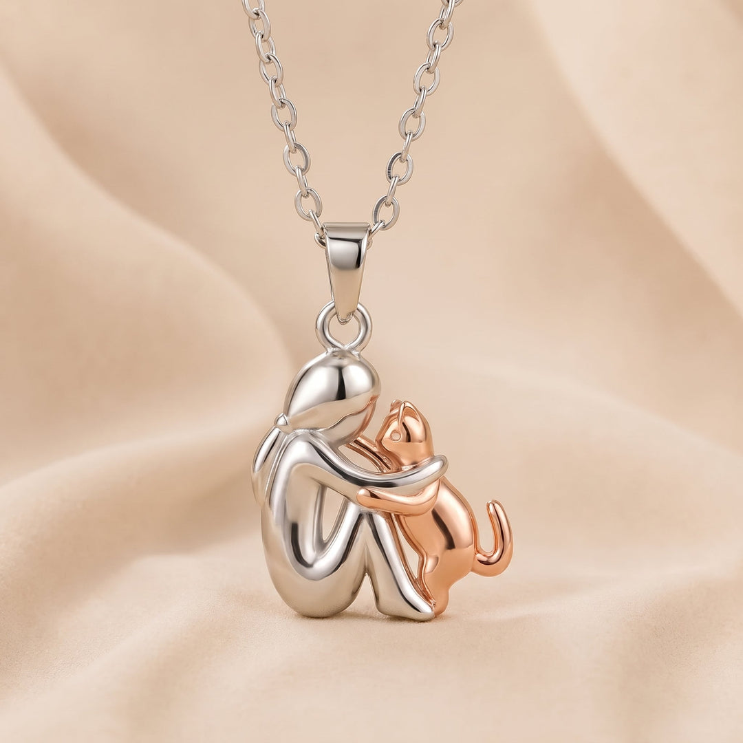 Silver-tone figure pendant with a rose-gold-tone cat reaching up to hug her, on a chain over peach fabric. 4