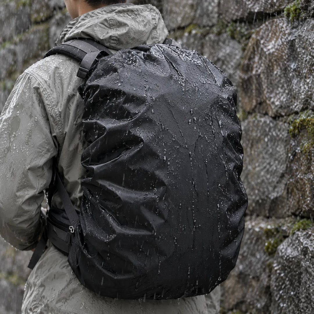 Hiker in a gray jacket seen from behind carries a black rain cover over a rucksack, water beading in front of a wet stone wall. 1