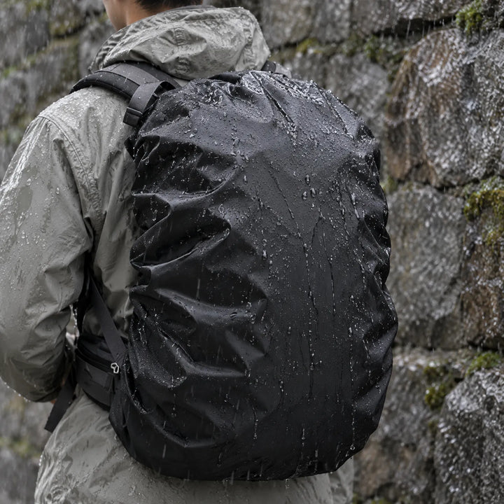 Hiker in a gray jacket seen from behind carries a black rain cover over a rucksack, water beading in front of a wet stone wall. 1