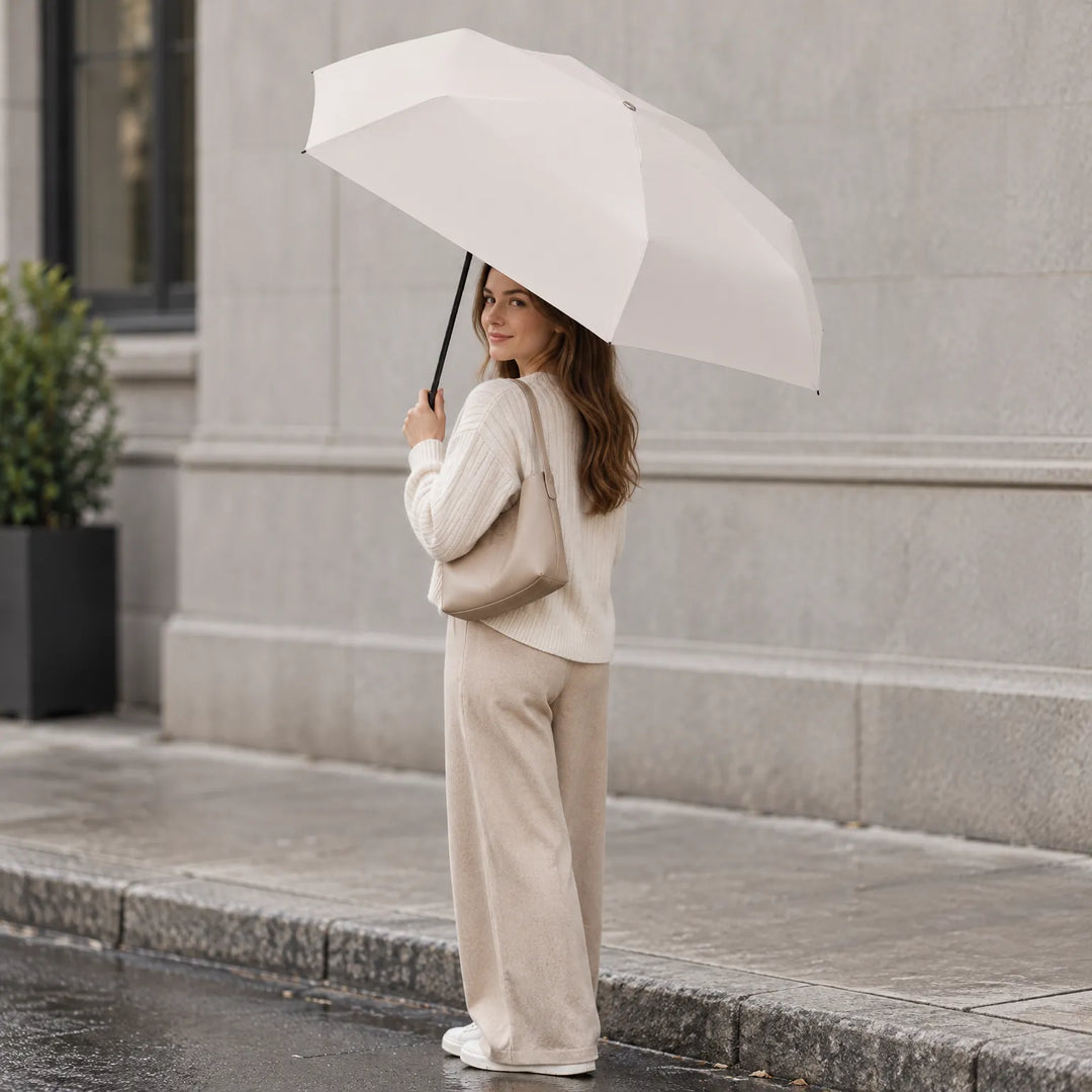 Woman in a cream sweater and beige trousers holds the open cream umbrella on a wet city sidewalk, shoulder bag on her arm. 1