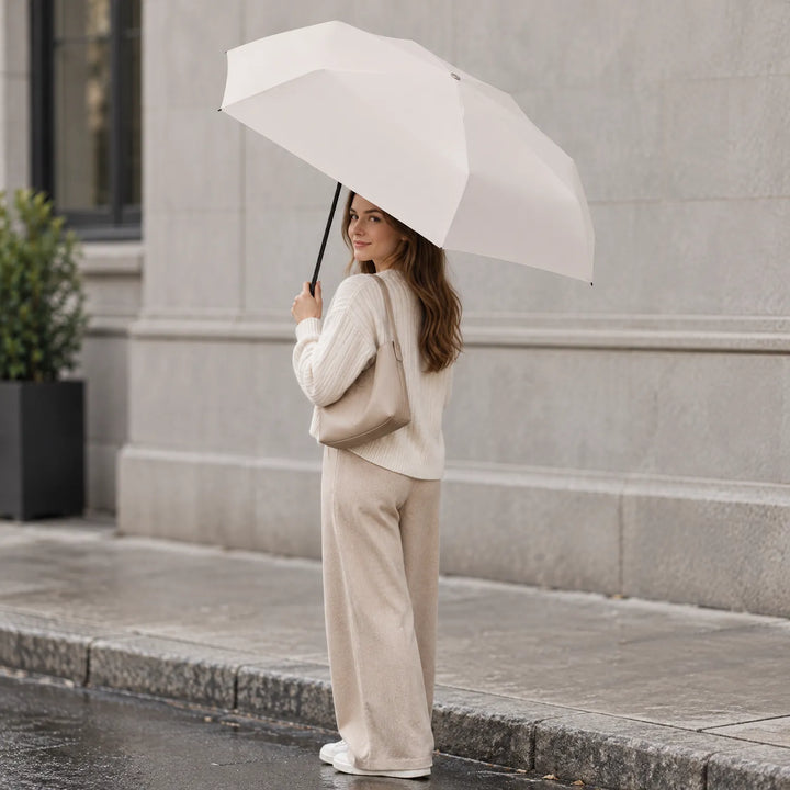 Woman in a cream sweater and beige trousers holds the open cream umbrella on a wet city sidewalk, shoulder bag on her arm. 1