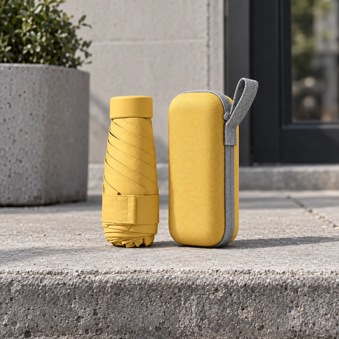Folded yellow umbrella and matching hard zipper case with gray loop standing on a concrete step beside a planter. 3