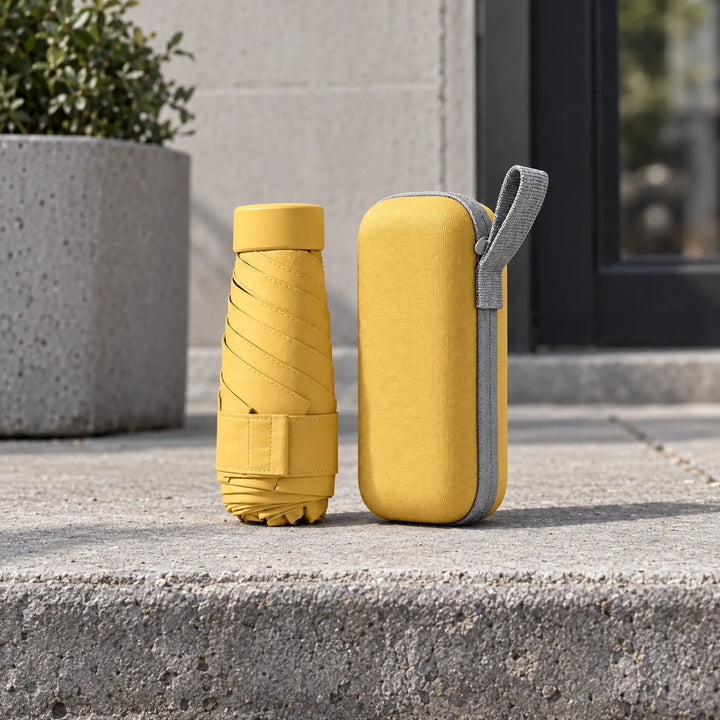 Folded yellow umbrella and matching hard zipper case with gray loop standing on a concrete step beside a planter. 3
