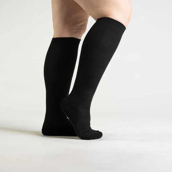 Side view of a model wearing black knee-high ribbed socks, one heel lifted to show the textured cushioned sole. 1