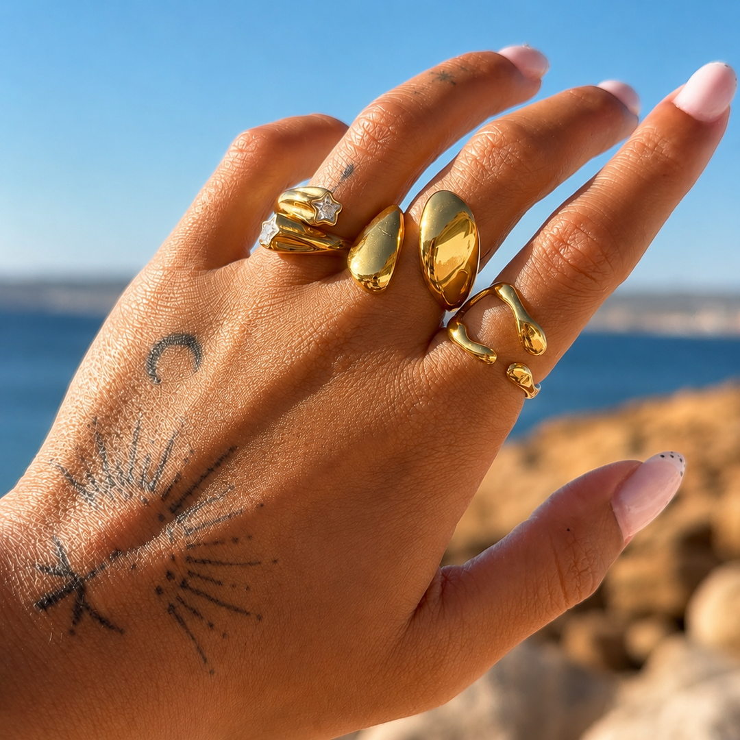 Hand with tattoos against a blue sea backdrop wearing the wavy gold open ring on one finger, stacked with other gold rings. 3