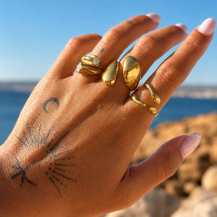Hand with tattoos against a blue sea backdrop wearing the wavy gold open ring on one finger, stacked with other gold rings. 3