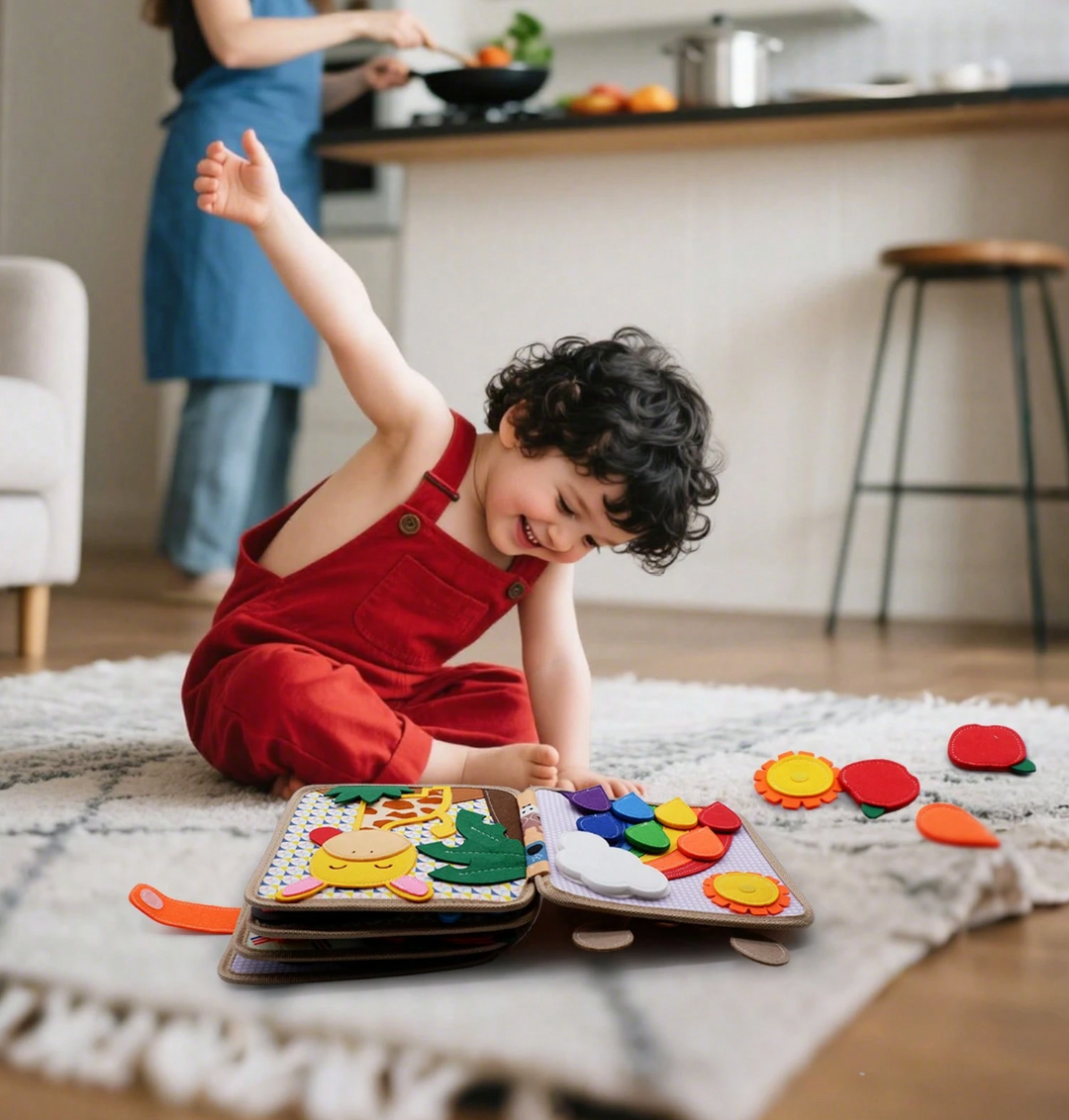 Laughing toddler in red overalls sitting on a rug beside the open felt book and loose felt pieces while a parent cooks.