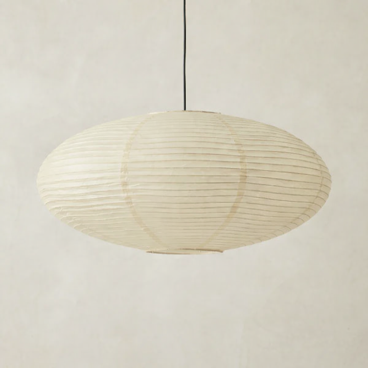 Oval cream paper pendant shade with horizontal ribs hanging from a black cord against a soft neutral wall. 1