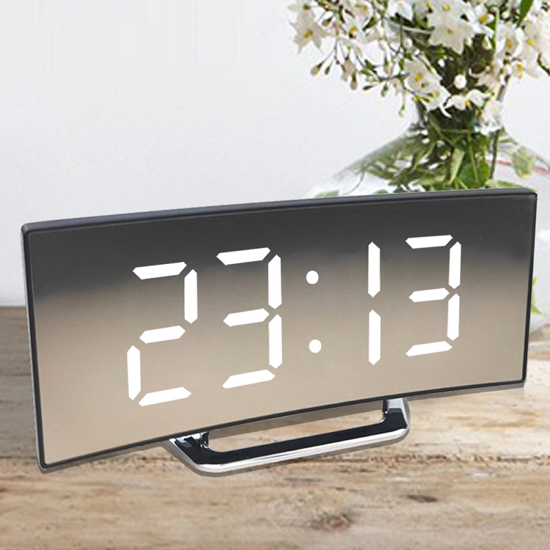 Curved mirror-front clock with large white digits on a wooden surface beside a vase of white flowers.