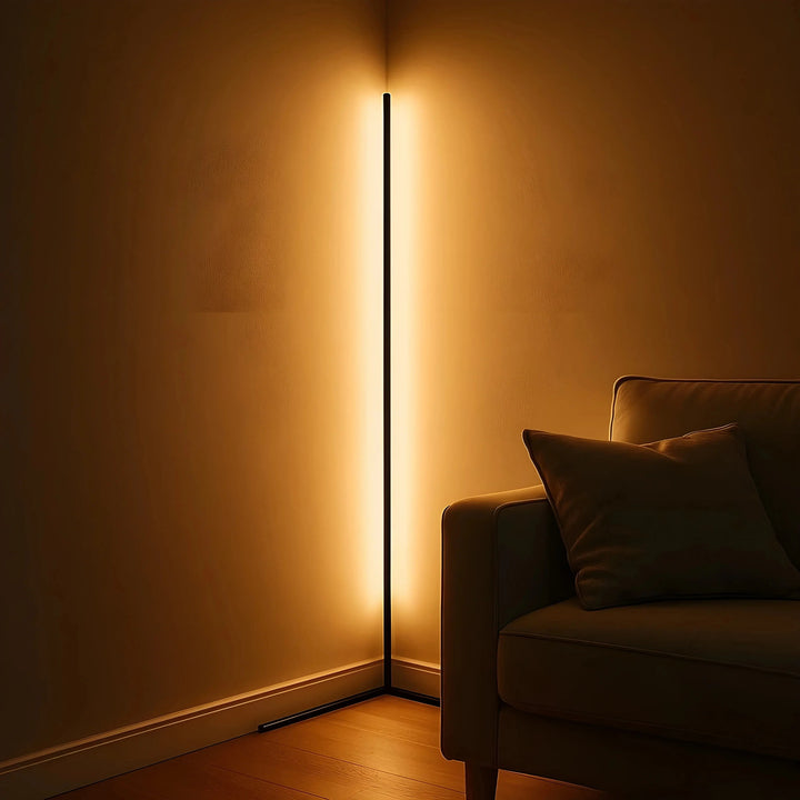 Black corner lamp lit in a deep amber glow in a dim room corner, with a sofa and cushion at right. 3