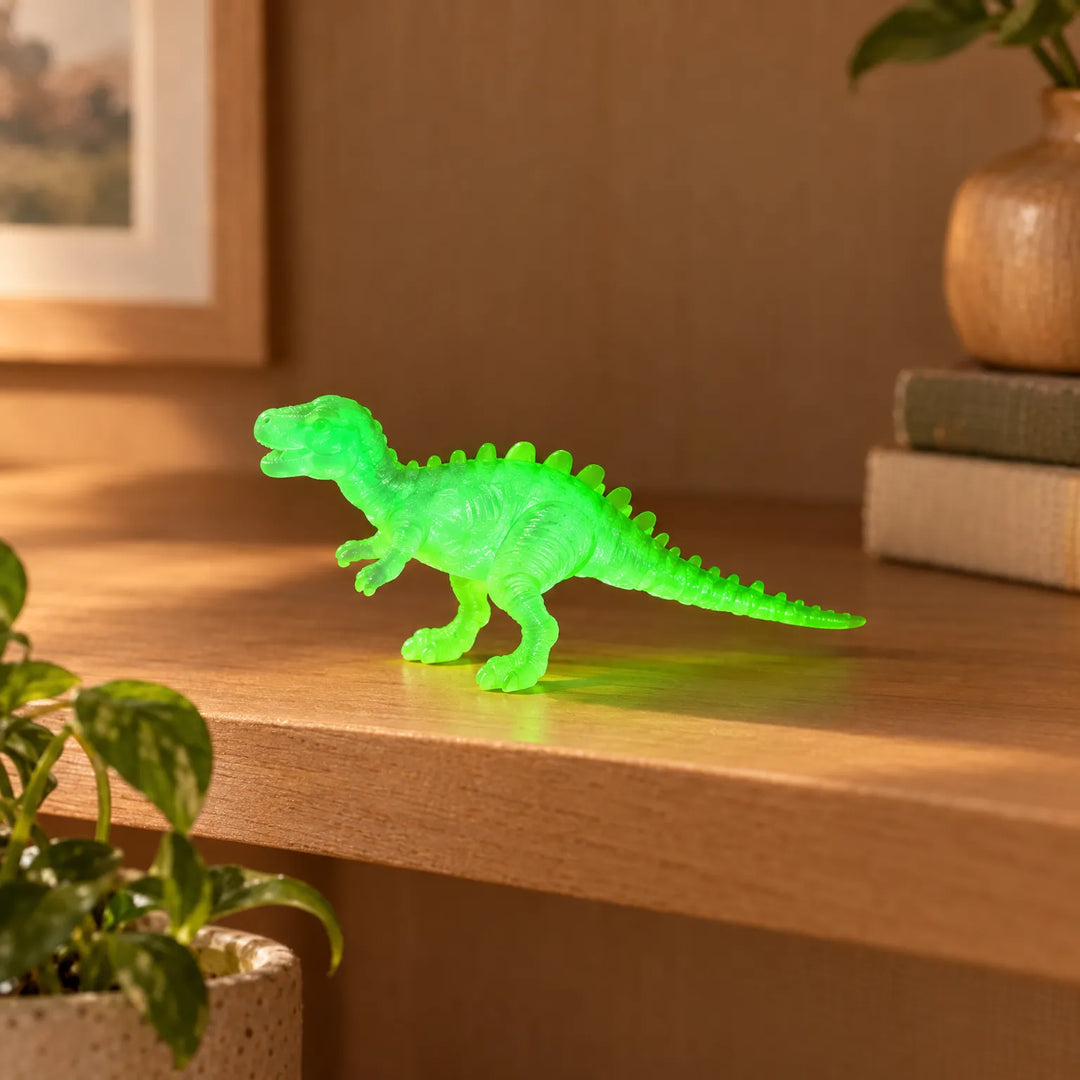 Single translucent bright green dinosaur figure standing on a wooden shelf beside books and a houseplant, with a soft green glow. 3