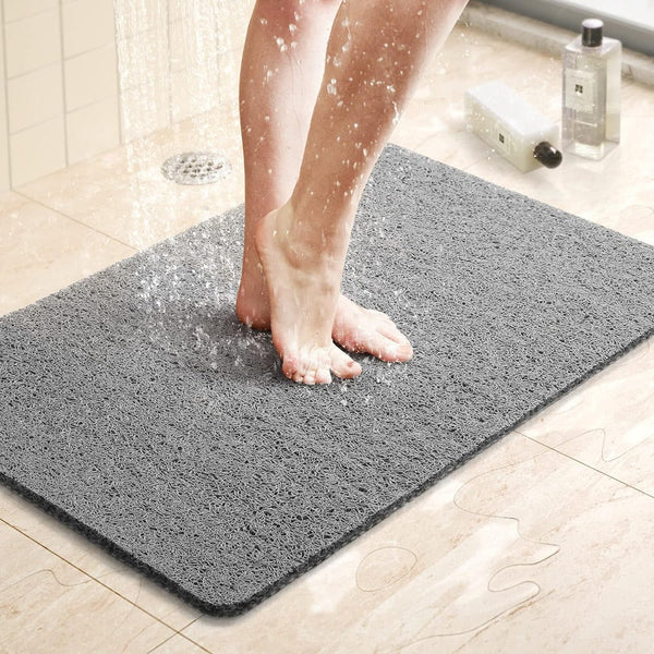Person's wet bare feet standing on a gray looped mat inside a shower, with water droplets splashing and a floor drain behind. 3