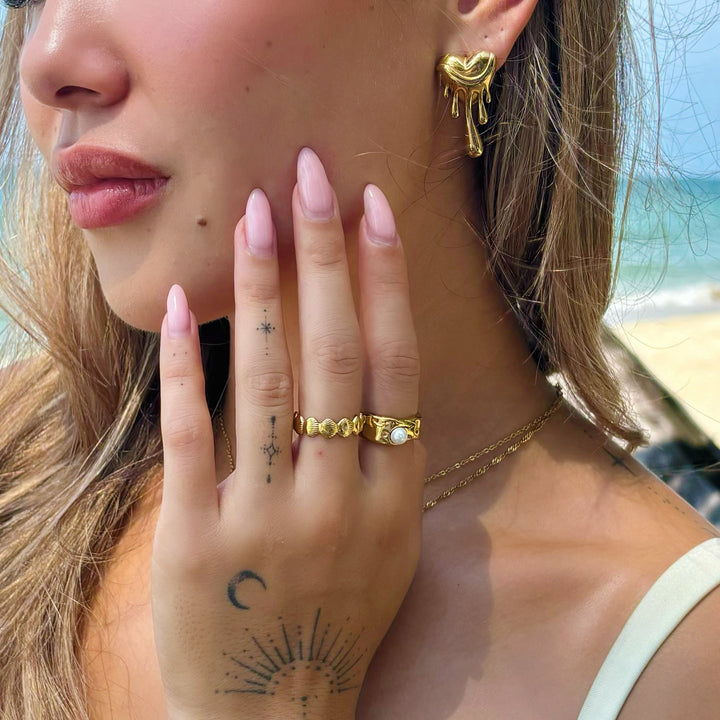 Close-up of a woman wearing one gold dripping heart earring, with gold rings and beach behind 4